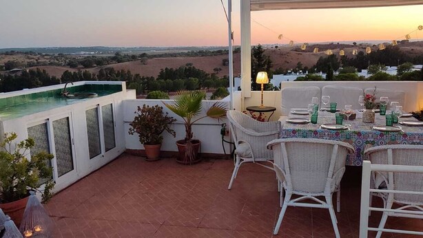Eine Terrasse mit Esstisch, gedeckt für vier Personen, umgeben von Korbstühlen, Topfpflanzen und Blick auf die Landschaft bei Sonnenuntergang.