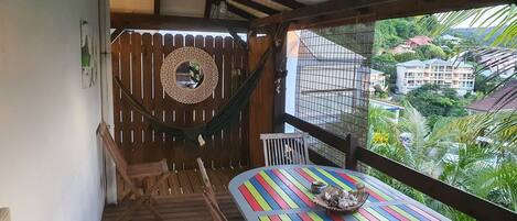 Un balcón con una hamaca, sillas de madera, una mesa colorida y vistas a edificios y follaje.