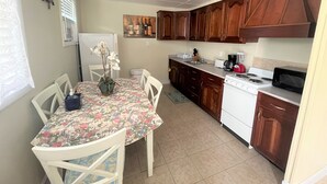 Een kleine keuken met houten kastjes, een witte oven en een magnetron. Er staat een eettafel met stoelen en een vaas met bloemen.