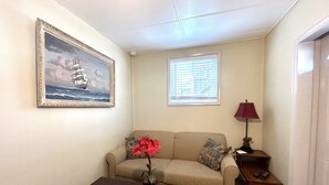 Een woonkamer met een beige bank, een klein tafeltje met een bloevas, een lijst met een schip erop, een raam met jaloezieën en een lamp op een bijzettafeltje.