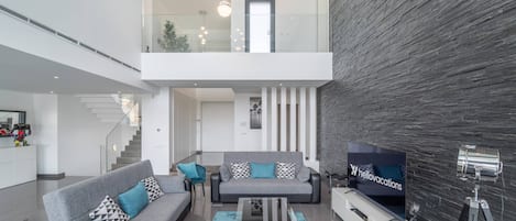 Un salon moderne avec un canapé gris, des coussins turquoise et une table basse en verre. Un téléviseur à écran plat est fixé au mur, et un trépied se trouve à proximité.