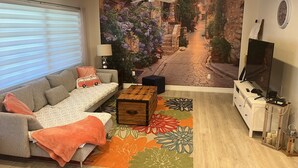 Una sala de estar con un sofá, una mesa de centro, un televisor y un mural que representa una escena callejera.