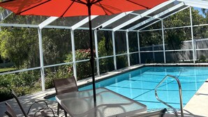 Zona de piscina exterior techada con mesa y sillas de vidrio, un paraguas rojo, y vistas a árboles y follaje.