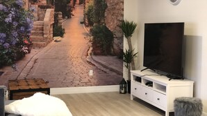 Una habitación con un televisor grande sobre un mueble blanco, un sofá acogedor con una manta blanca y un mural de una calle adoquinada.