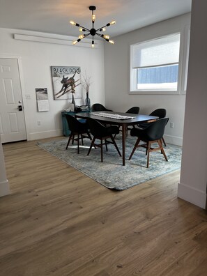 A dining area with a wooden table, black chairs, a blue rug, a ceiling light fixture, and a window with blinds.
