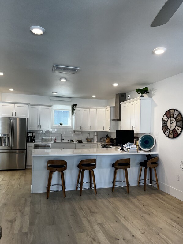 A modern kitchen with white cabinets, a stainless steel refrigerator, a large island with bar stools, and a wall-mounted clock.