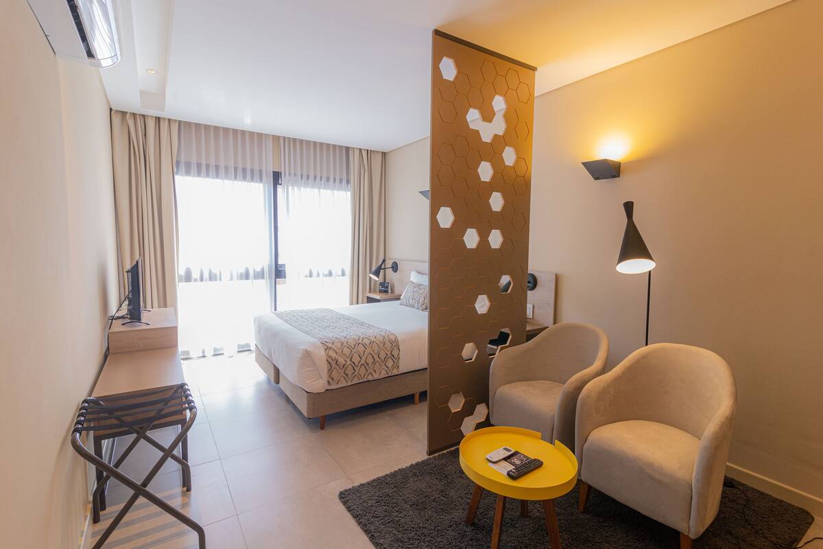 Habitación de hotel con cama, escritorio, silla, mesita, lámpara y vista a la ciudad desde la ventana.
