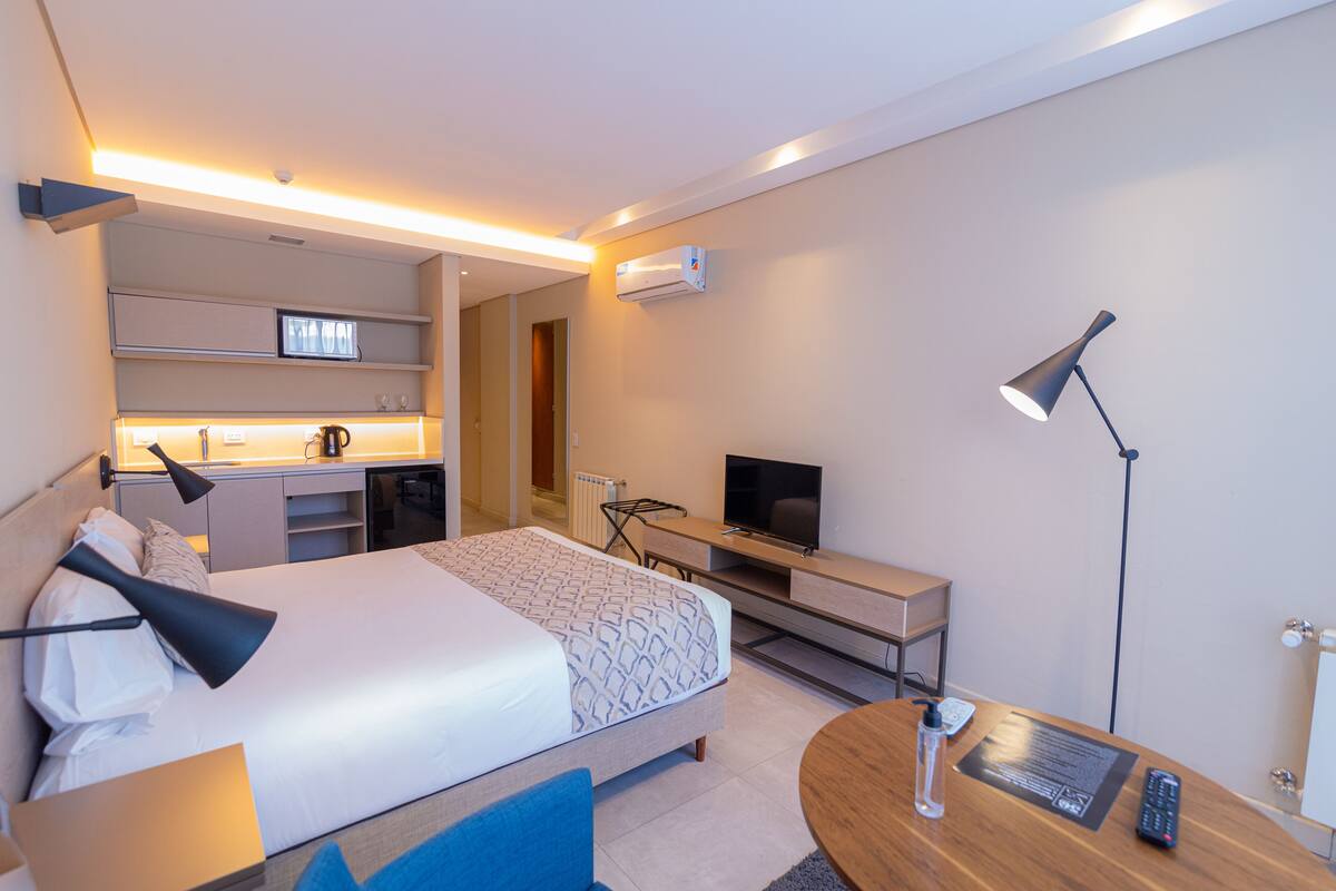 Una habitación de hotel moderna con cama, mesita de noche, televisor, escritorio y kitchenette.