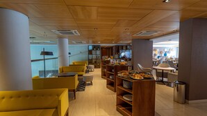 Un café moderne avec des chaises jaunes, des étagères en bois où sont exposés des produits alimentaires, et un téléviseur en arrière-plan.