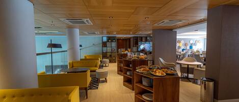 A modern cafe with yellow chairs, wooden shelves with food, and a TV in the background.
