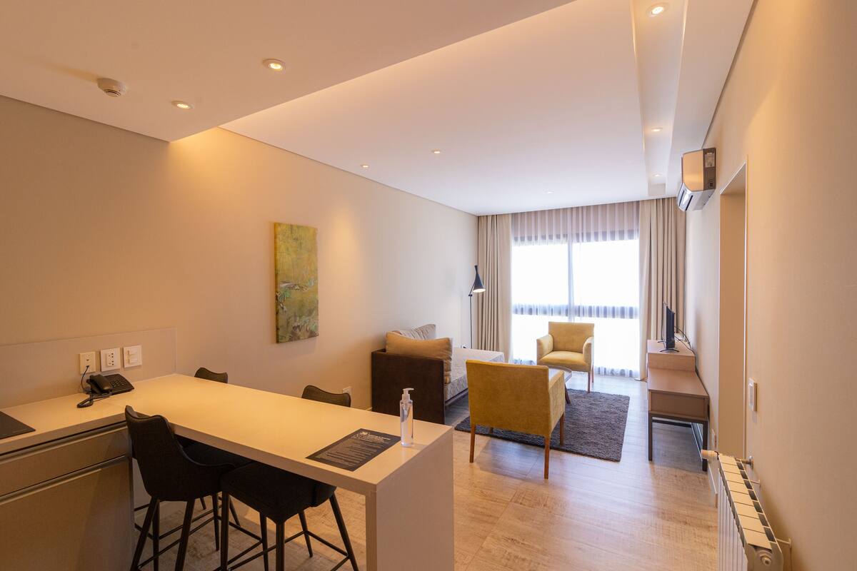 Una habitación de hotel moderna con kitchenette, comedor y sala de estar con sofá y sillones.