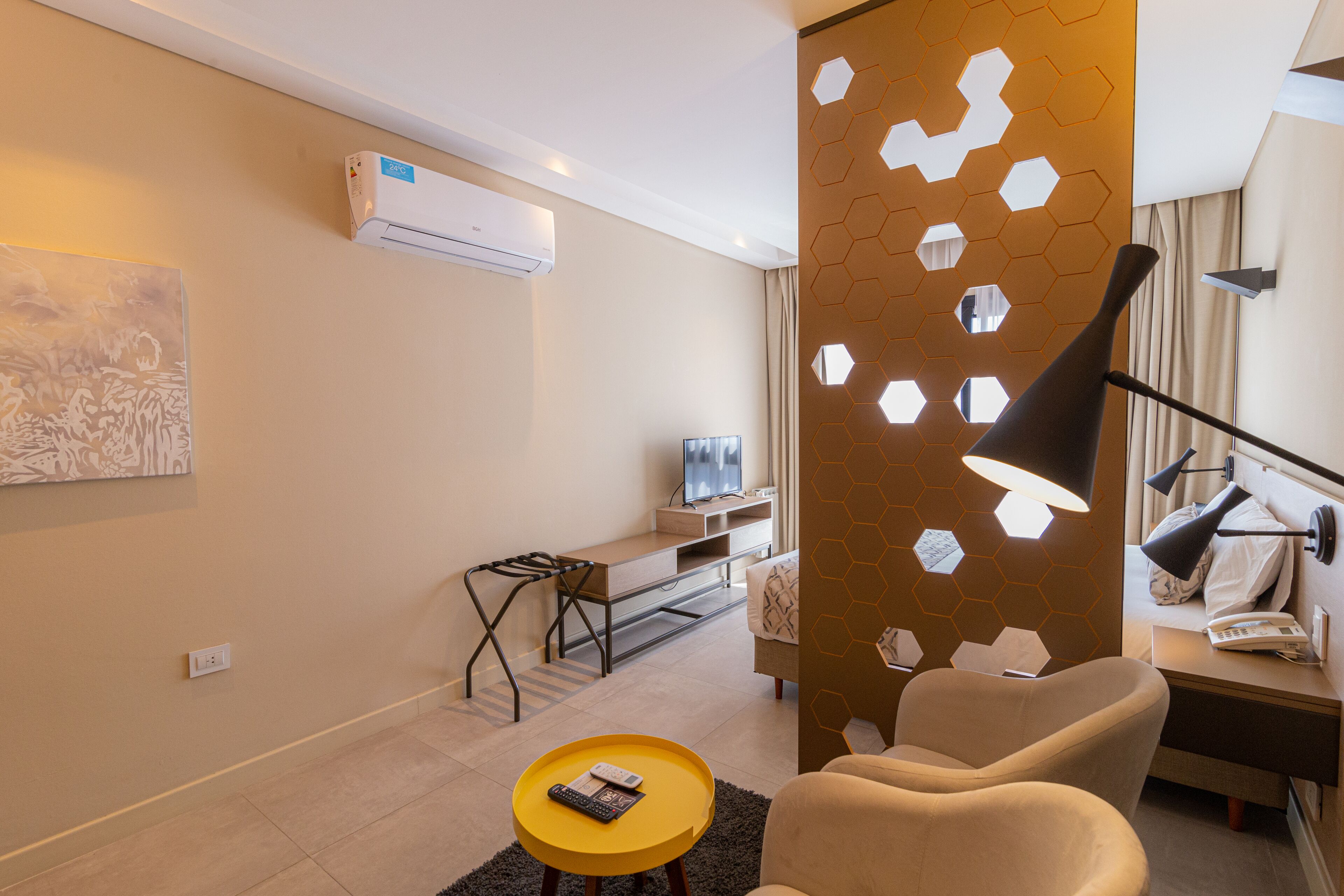 A modern hotel room with a flat-screen TV, a desk, a chair, a sofa, and a decorative wall with hexagonal cutouts.