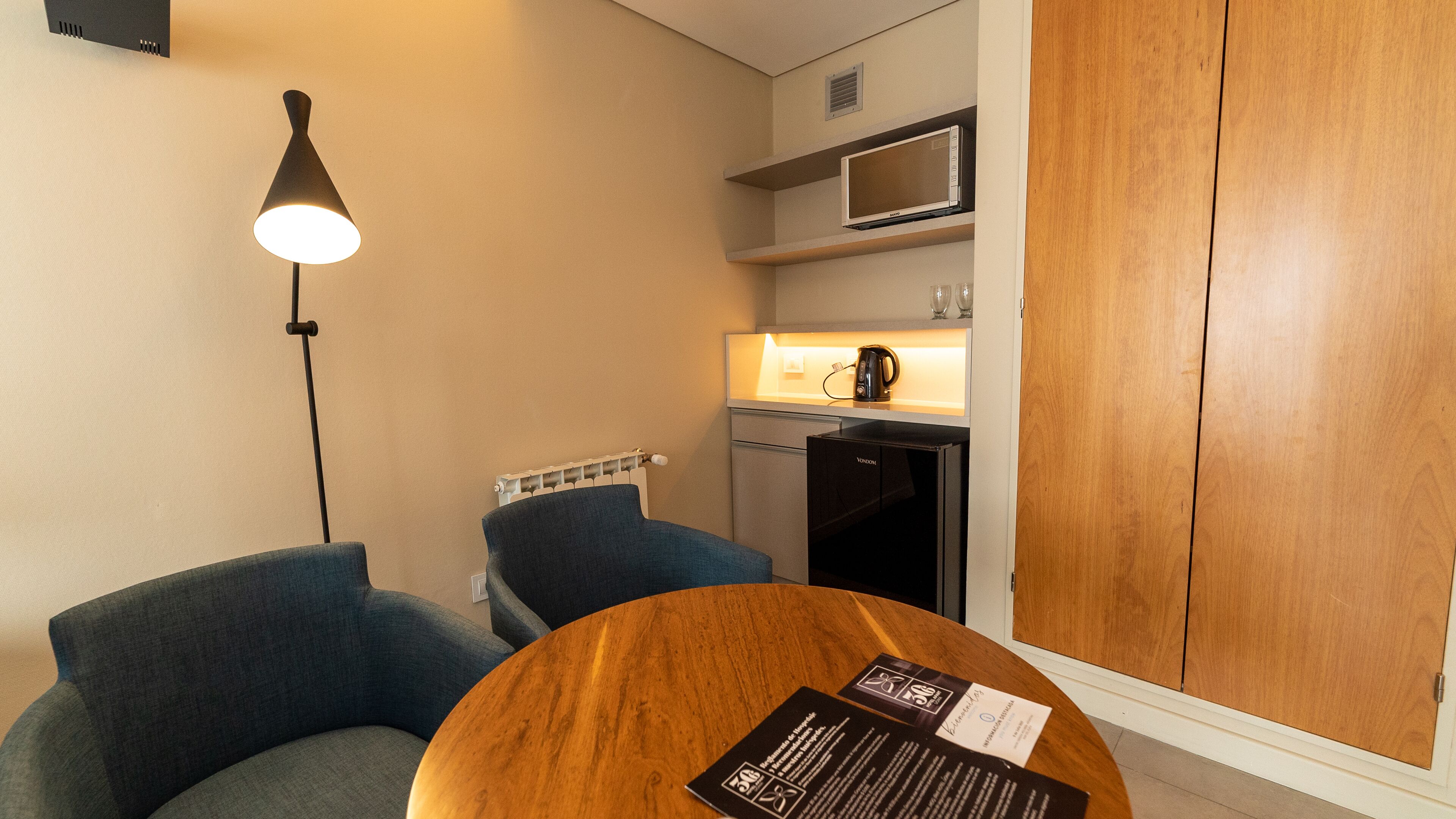 A compact hotel room with a kitchenette, a round wooden table, a blue armchair, and a standing lamp.