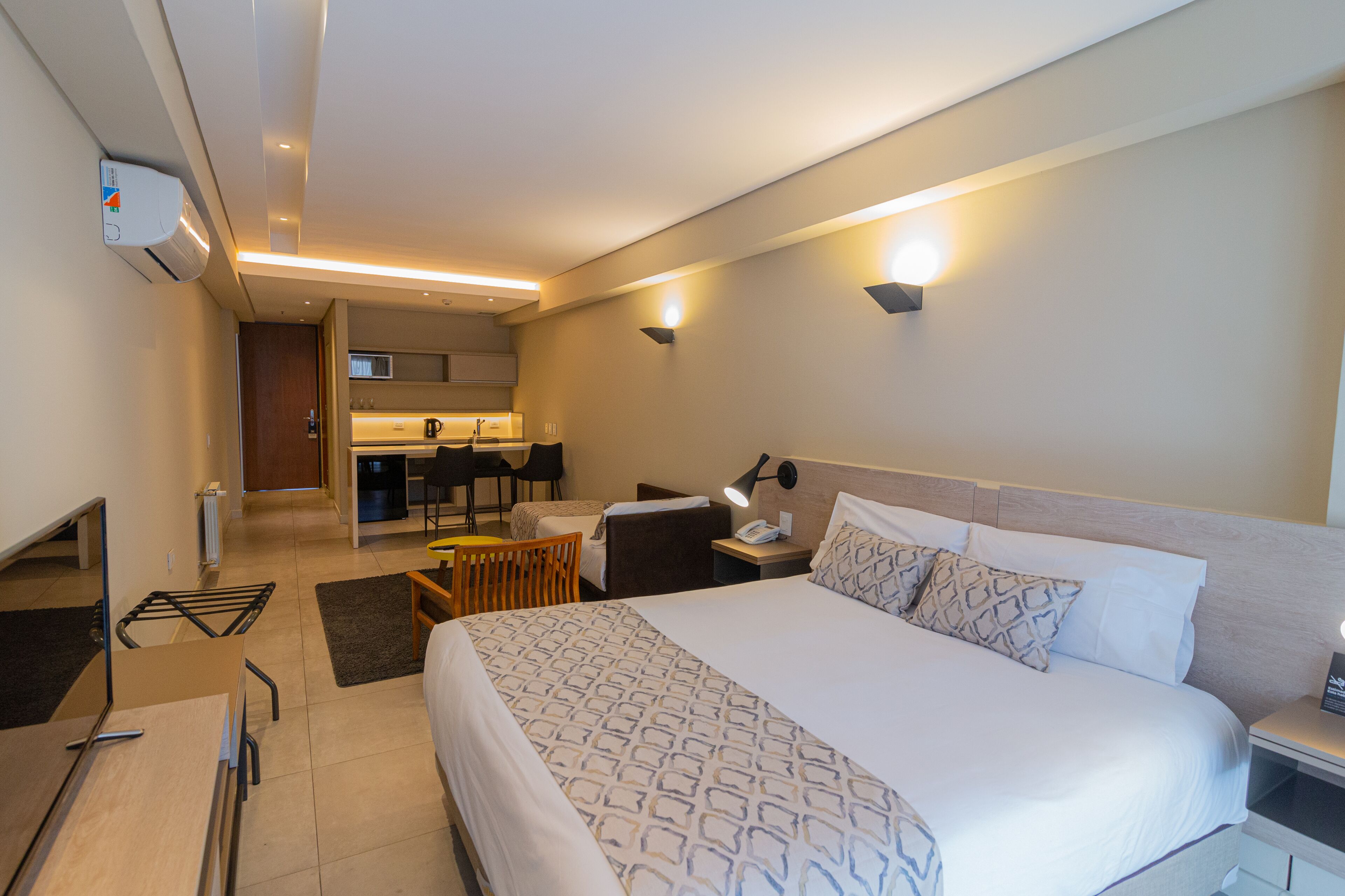 Habitación de hotel con una cama grande, mesita de noche y un escritorio con silla. Se ve una kitchenette al fondo.