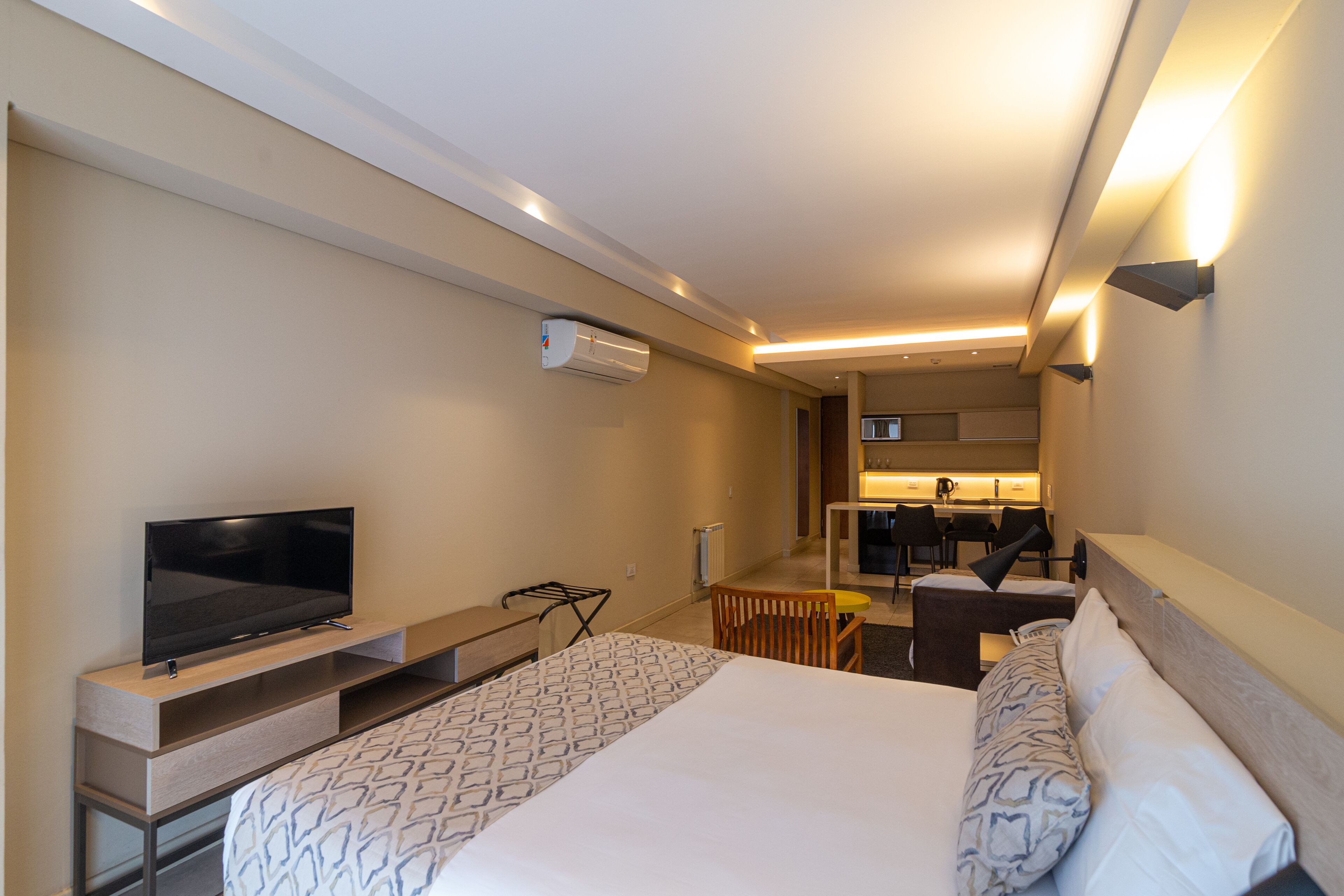 A modern hotel room with a large bed, a flat-screen TV, a small dining table, and a kitchenette in the background.