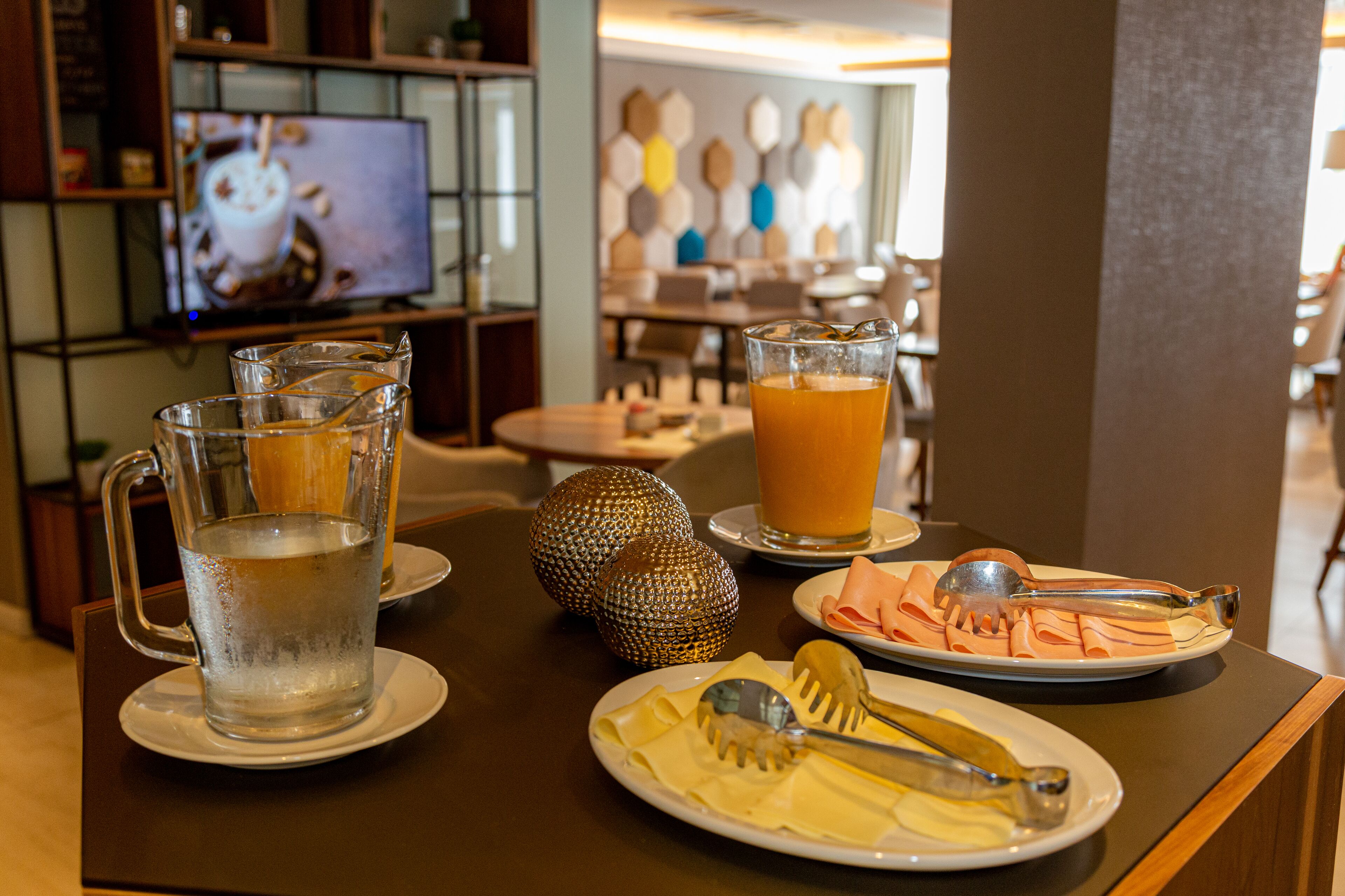 A dining table with a pitcher of water, a glass of orange juice, and plates of food including cheese and ham. A television is in the background displaying a beverage.