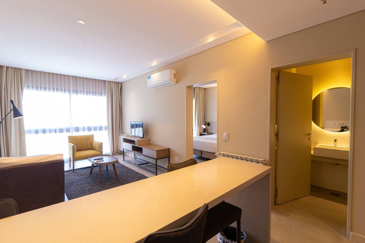 Habitación de hotel con una cama grande, un escritorio, una silla, un televisor, una lámpara y un baño con espejo y lavamanos.