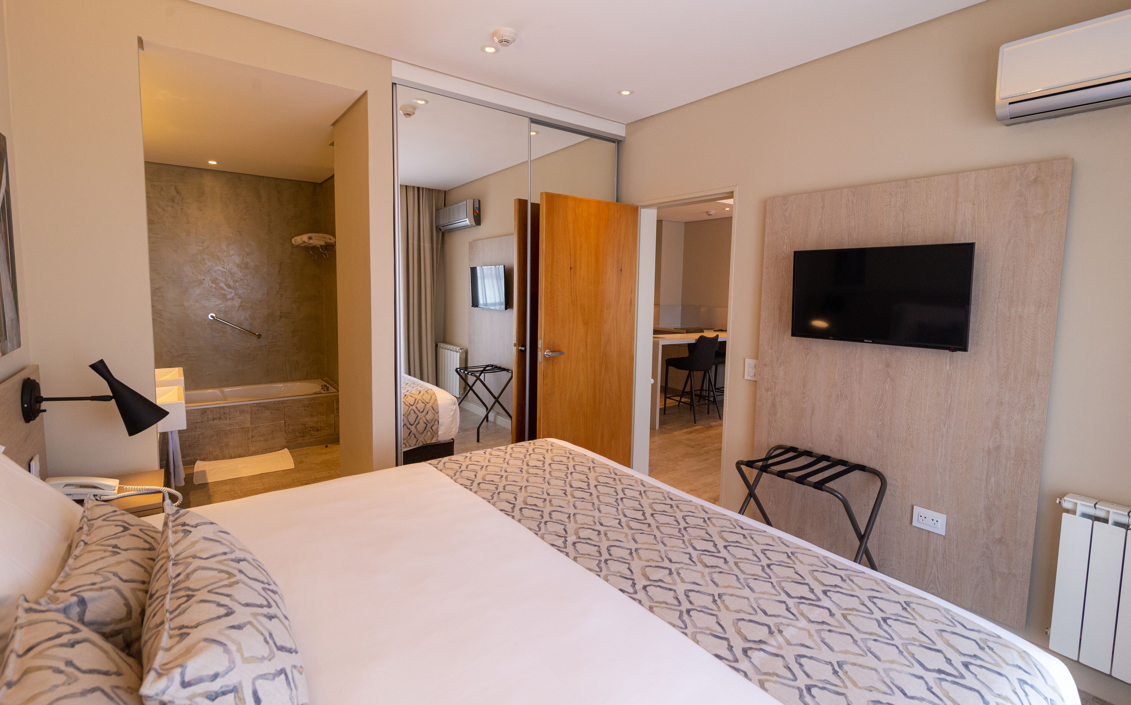 Una habitación de hotel moderna con una cama grande, un televisor montado en la pared, zona de ducha y un pequeño rincón de descanso con una silla.