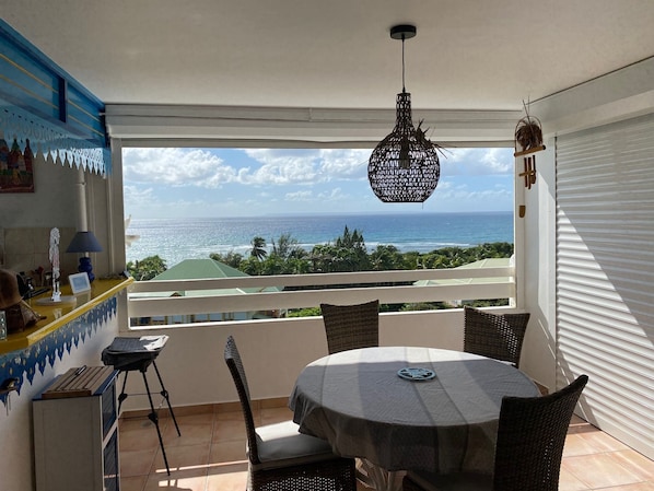 A balcony with a view of the ocean, a round table, wicker chairs, a hanging light fixture, and a counter with a lamp.