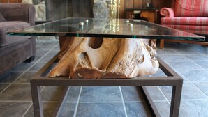 Een glazen salontafel met houten poten, geplaatst op een betegelde vloer in een kamer met een stenen muur en een ruitjesbank.