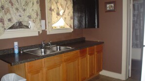 A kitchen with wooden cabinets, a double sink, and a window with floral curtains. - The Little Sunflower (Jetmore)