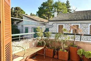 A balcony with a metal chair, potted plants, and a view of a stone building with a tiled roof.