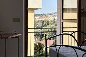 A room with a bed, a small table, and a balcony with a view of buildings and trees.