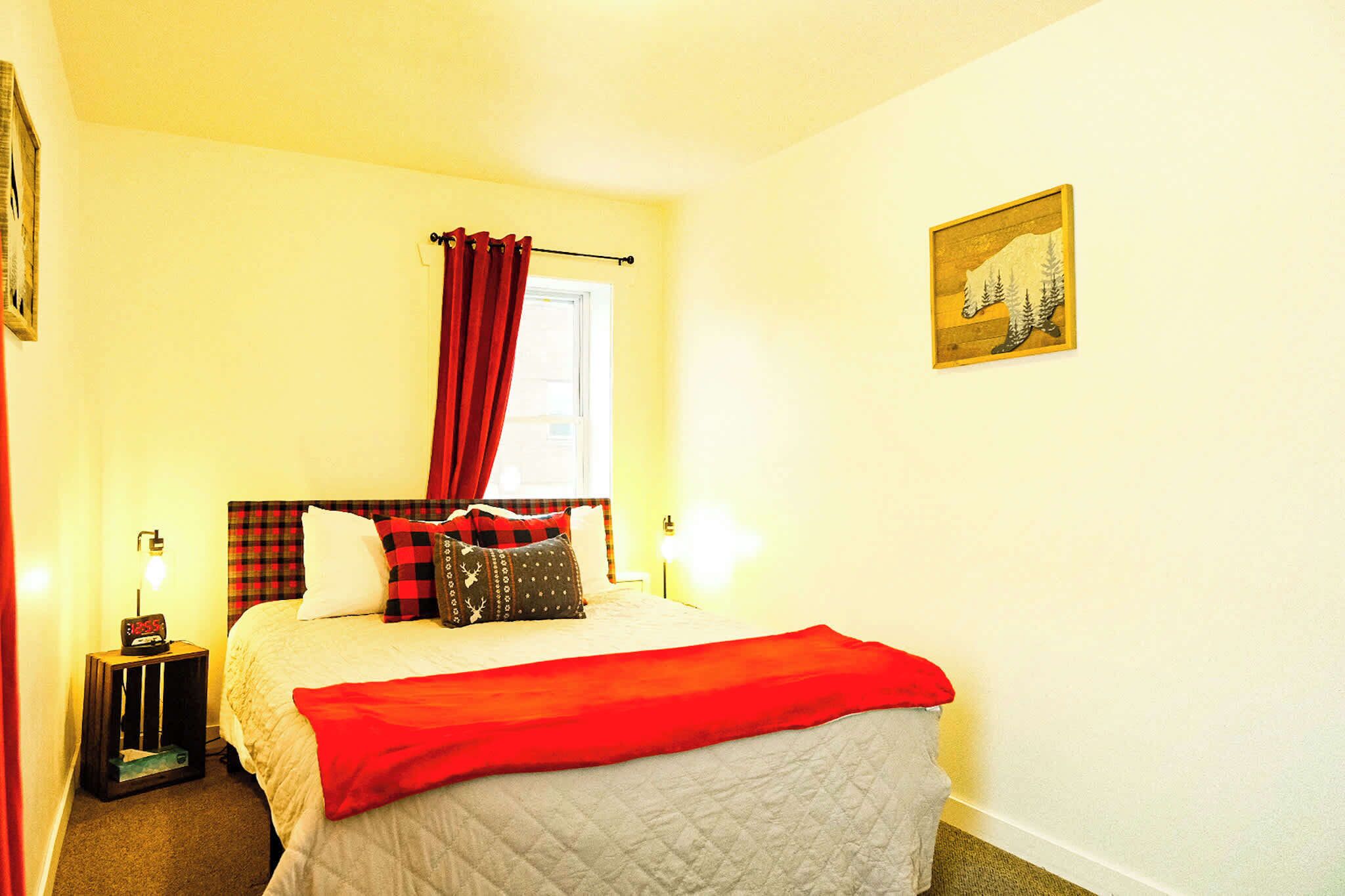 A bed with a red blanket, a red and black checkered headboard, and a window with red curtains.
