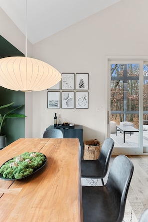 Uma sala de jantar moderna com mesa de madeira, cadeiras pretas e um pendente. Há quadros nas paredes e vista para o exterior através de uma porta de vidro.