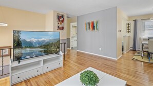 Een moderne woonkamer met een flatscreen televisie, houten vloeren, een witte salontafel en een eethoek met een tafel en stoelen.