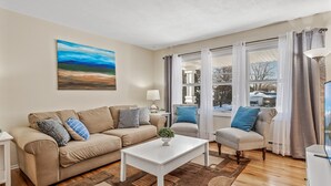 Een woonkamer met een beige bank, een witte salontafel en zicht op een besneeerd landschap door grote ramen.