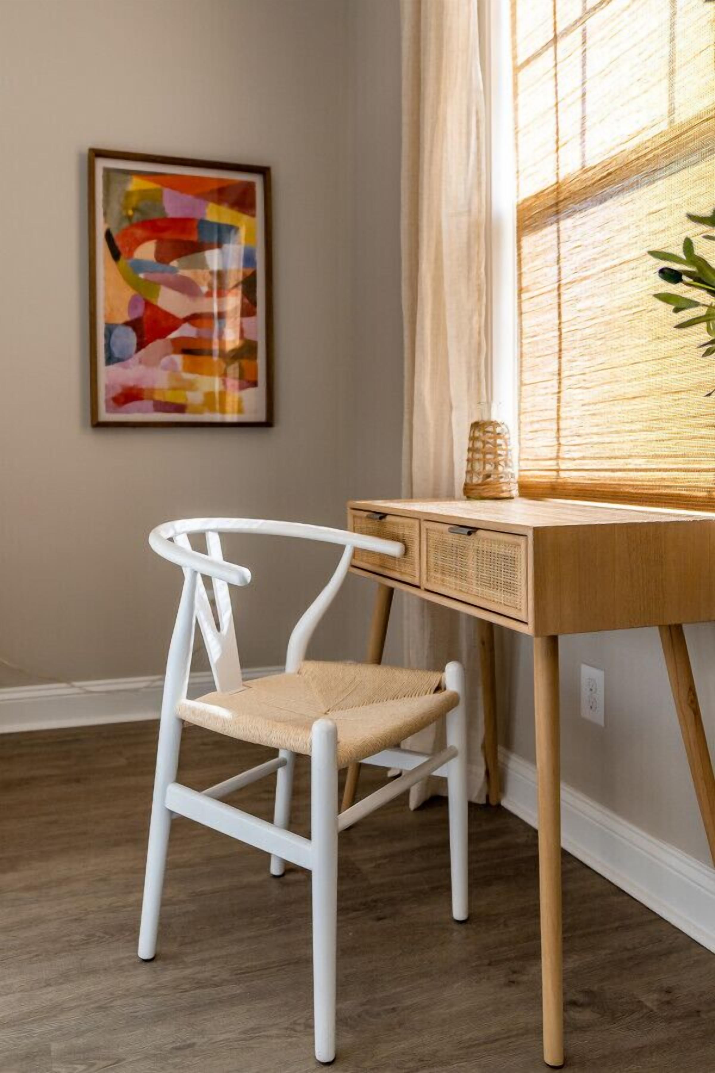 Una sedia bianca con seduta intrecciata, una scrivania di legno con un cesto di vimini e un quadro astratto dai colori vivaci appeso alla parete.