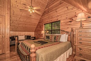 Quarto de cabana de tronco com cama, penteadeira, mesa de cabeceira, ventilador de teto e janela com persianas.