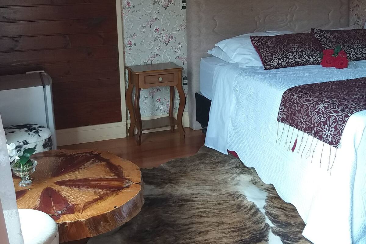 Une chambre avec un plancher en bois, un tapis en peau de vache, un lit avec une literie blanche et à motifs, une petite table de chevet en bois, un lustre et un papier peint à motifs floraux.