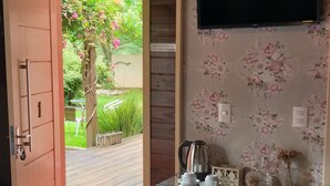 A room with a wooden door leading to a garden, a television mounted on the wall, a cabinet with kitchen items, and a bed with a patterned bedspread. - POUSADA REQUINTE SERRANO (Urubici)