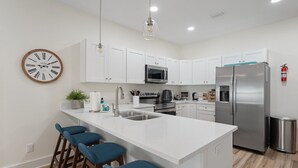 Una cocina moderna con gabinetes blancos, un refrigerador de acero inoxidable, un microondas y un lavamanos. Hay taburetes altos azules y un reloj en la pared.