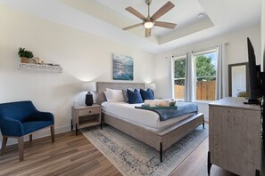 Ein Schlafzimmer mit einem Bett, einem blauen Sessel, einem Nachttisch, einem Schreibtisch, einem Deckenventilator und einem Fenster mit Vorhängen.