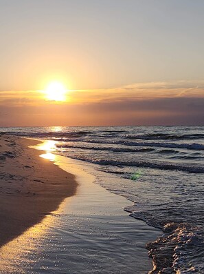 Ein Strand bei Sonnenuntergang, die Sonne tief am Horizont, ihr Spiegelbild im Wasser.