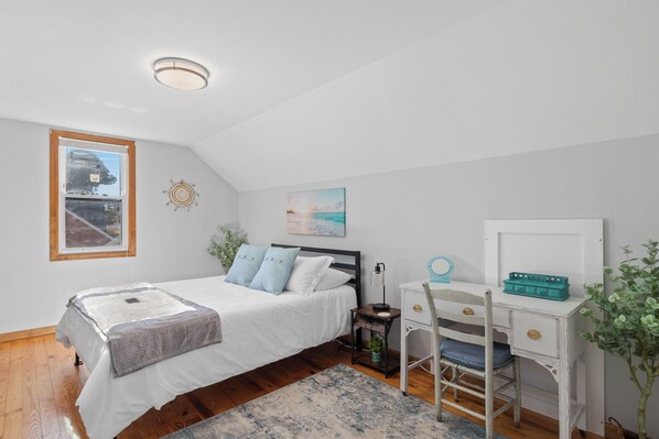 A bedroom with a bed, a desk, a chair, a small table, and a window.
