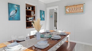 Un coin repas avec une table en bois dressée pour six personnes, des murs blancs et des œuvres d’art représentant des oiseaux et des poissons.