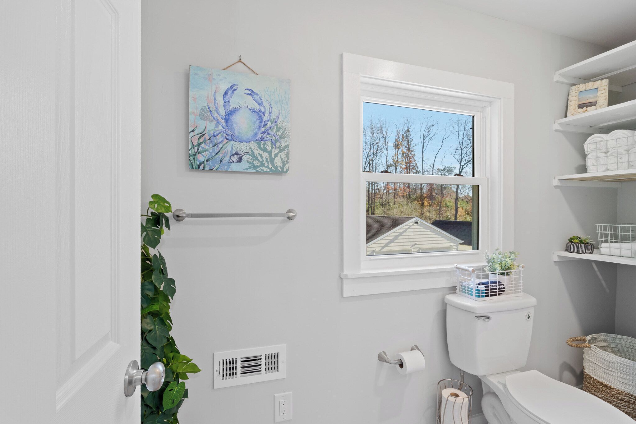 A bathroom with a toilet, a window with a view of trees, a towel rack, and a painting on the wall.