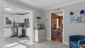 Une cuisine moderne avec des meubles blancs, des appareils en acier inoxydable et un meuble en bois abritant une machine à café. On y trouve également un coin repas avec un canapé bleu et un tableau accroché au mur.