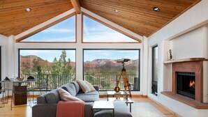 Un salón amplio con una ventana grande que da a un paisaje montañoso, una chimenea y un telescopio sobre un trípode.
