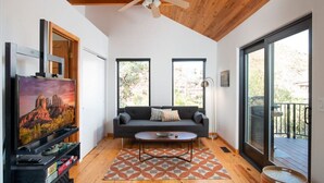 Una sala de estar moderna con un sofá gris, una mesa de centro y un televisor sobre un mueble bajo. Se ven ventanas grandes y un balcón.