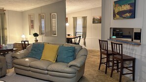 En stue med en beige sofa, træstole, et spisebord og et køkkenområde med en mikrobølgeovn.