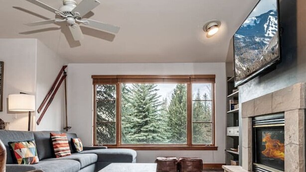 A modern living room with a fireplace, a ceiling fan, and a large window overlooking a forest. - Luxurious Condo Gated Community Pool Hot Tubs (Edwards)