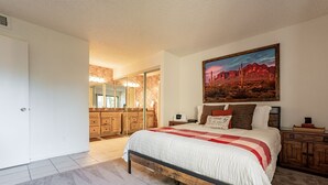 A bedroom with a large bed, a nightstand, a dresser, and a painting of a desert landscape.