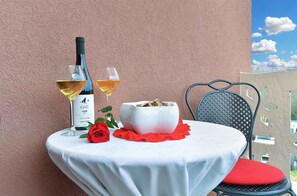 A table with a white tablecloth, two wine glasses, a bottle of wine, a white bowl with food, a red rose, and a red cushion on a chair.