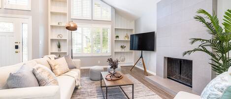 A modern living room with a sofa, coffee table, and TV. There is a fireplace and a large window with blinds.