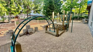 Ein Spielplatz mit Holzkonstruktionen und Metallrahmen, umgeben von Kies und Bäumen.